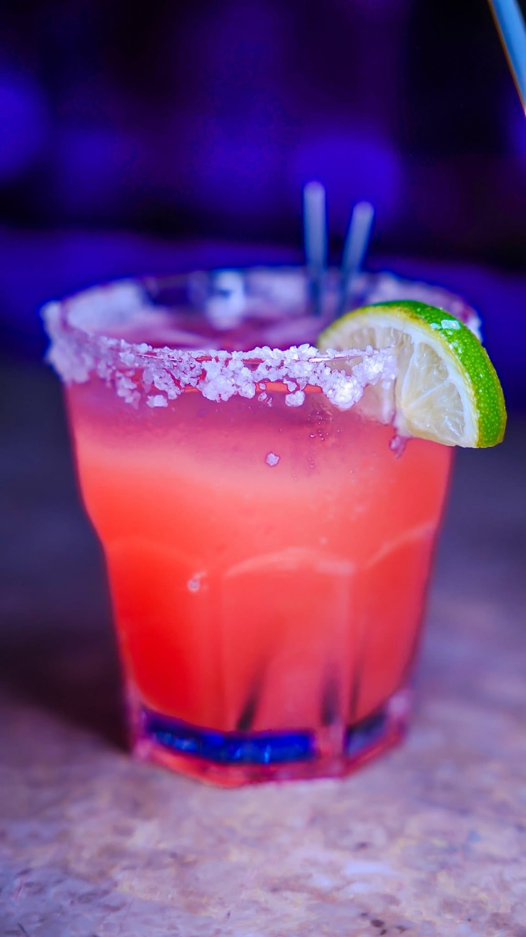 Salt-rimmed strawberry margarita with a lime wheel under neon